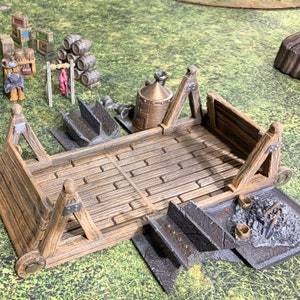 May include: A wooden siege tower with a wooden floor and metal wheels. The tower is on a green grassy surface. There are other miniature models in the background.