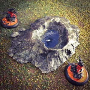 May include: A miniature model of a volcanic crater with a small pool of water at the bottom. The crater is made of grey and brown textured material. Two miniature figures of demons are standing on bases with orange and black flames.