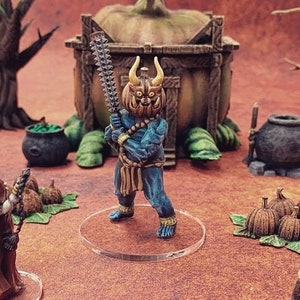 May include: A blue and gold painted miniature figure of a demon holding a sword. The figure is standing on a clear plastic base. The figure is surrounded by other miniature figures and objects, including a pumpkin, a cauldron, and a tree.