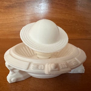 May include: White 3D printed model of a planet with rings, sitting on a white base with a decorative design.