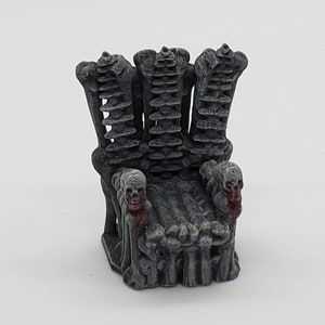 May include: A miniature skeletal throne made of gray plastic with two skulls on the armrests. The skulls have red paint on the mouth area.