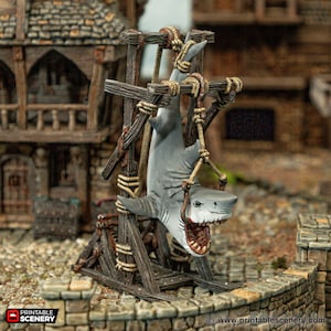 May include: A wooden structure with a rope and pulley system holds a hanging shark with its mouth open. The shark is painted gray and has a white underbelly. The structure is made of weathered wood and has a rustic appearance.
