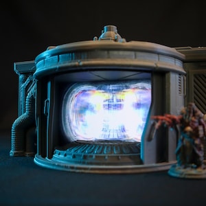 May include: A grey, futuristic-looking structure with a glowing blue and white portal in the centre. The structure has a detailed design with pipes and wires visible.