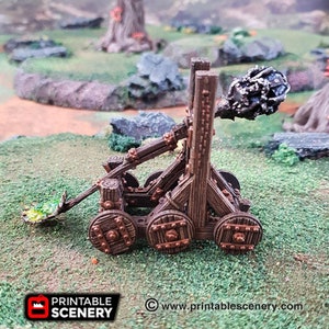 May include: A wooden catapult with a metal projectile, painted in brown and copper tones. The catapult is on a green grassy surface. The text "www.printablescenery.com" is visible in the lower right corner of the image.