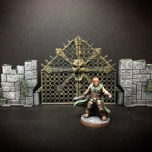 May include: A miniature figure of a warrior standing in front of a detailed, intricately designed, metal gate. The gate is made of a dark metal with a honeycomb pattern and is flanked by two stone walls. The warrior is wearing a green cloak and holding a sword.