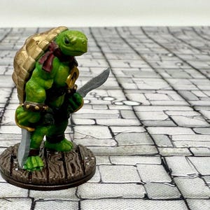 May include: A green and gold painted miniature turtle warrior figurine holding a silver sword. The figurine is standing on a wooden base.