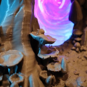 Peut inclure: Une sc&egrave;ne miniature imprim&eacute;e en 3D avec un portail lumineux violet et bleu entour&eacute; de champignons et de rochers gris.