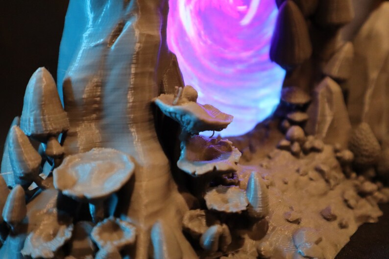 Peut inclure: Une sc&egrave;ne miniature imprim&eacute;e en 3D avec un portail lumineux violet et bleu entour&eacute; de champignons et de rochers gris.