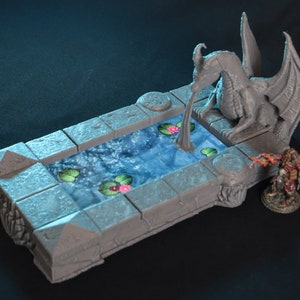 May include: A gray stone fountain with a dragon statue pouring water into a pool with blue water and pink flowers. The fountain is made of multiple pieces and has a detailed design.