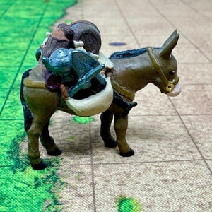 May include: A brown donkey figurine with a saddle and a small figure riding on its back. The donkey is carrying a variety of items, including a barrel, a bag, and a sword.