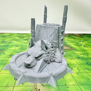 May include: A gray 3D printed miniature of a wolf head with a wooden structure behind it. The wolf head is on a circular base with spikes around the edge. The structure is made of logs and has a ladder on the side.