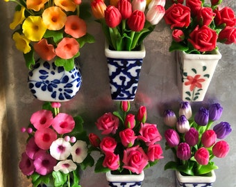 May include: A collection of colorful miniature flower arrangements in ceramic vases. The vases feature blue and white patterns, and the flowers include roses, tulips, and other blooms in various colors like pink, red, and blue. The arrangements are displayed on a metallic surface.