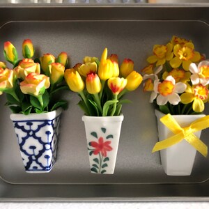 Op de afbeelding: Drie miniatuurbloemstukken in keramische potten. De eerste pot is blauw en wit met een bloemenpatroon, de tweede pot is wit met een rode bloem en groene bladeren, en de derde pot is wit met een gele strik. De bloemen zijn geel en wit.