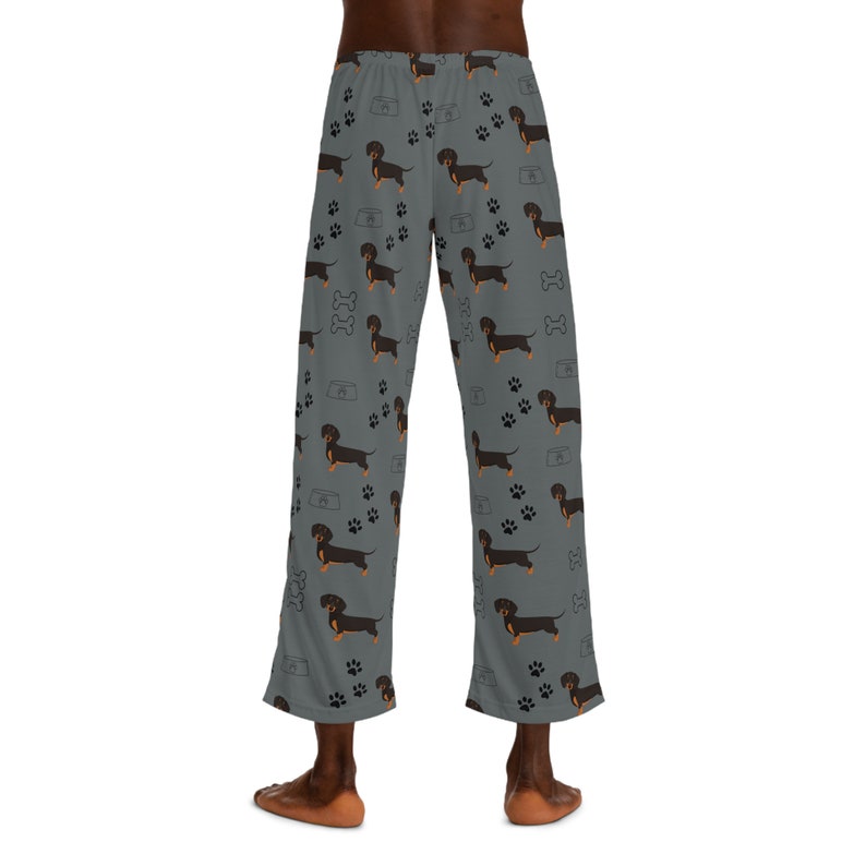 May include: Gray lounge pants featuring an all-over pattern of black and tan dachshund dogs, black paw prints, white dog bone outlines, and white food bowl outlines. The pants have a straight-leg cut and an elasticized waistband at the top.