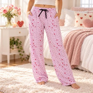 May include: Light pink pajama pants featuring a repeating pattern of dark gray dachshund dog outlines and small red hearts. The pants have a black drawstring at the elastic waistband and two side pockets, providing a relaxed fit.
