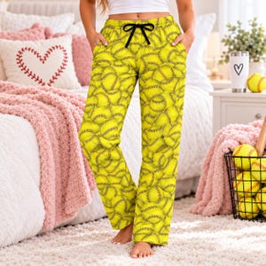 May include: Bright yellow lounge pants featuring an all-over print of softball patterns with red stitching. The pants include a black drawstring waistband and two side pockets, providing a relaxed fit for casual wear.