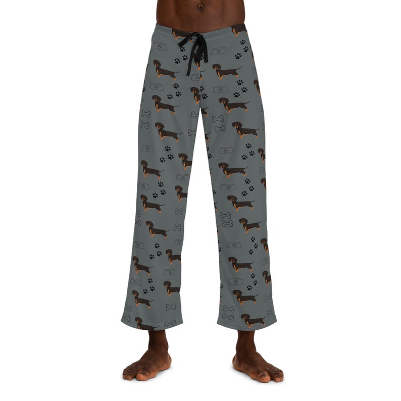May include: Grey lounge pants featuring a repeating pattern of black and tan dachshund dogs, black paw prints, white dog bones, and grey food bowls. The pants have a black drawstring at the elastic waistband and a straight-leg cut.