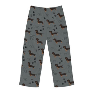 May include: Gray lounge pants featuring an all-over repeating pattern of black and tan dachshund dogs, black paw prints, white dog bones, and light gray dog bowls. The pants have a straight-leg cut and a simple elasticized waistband design.
