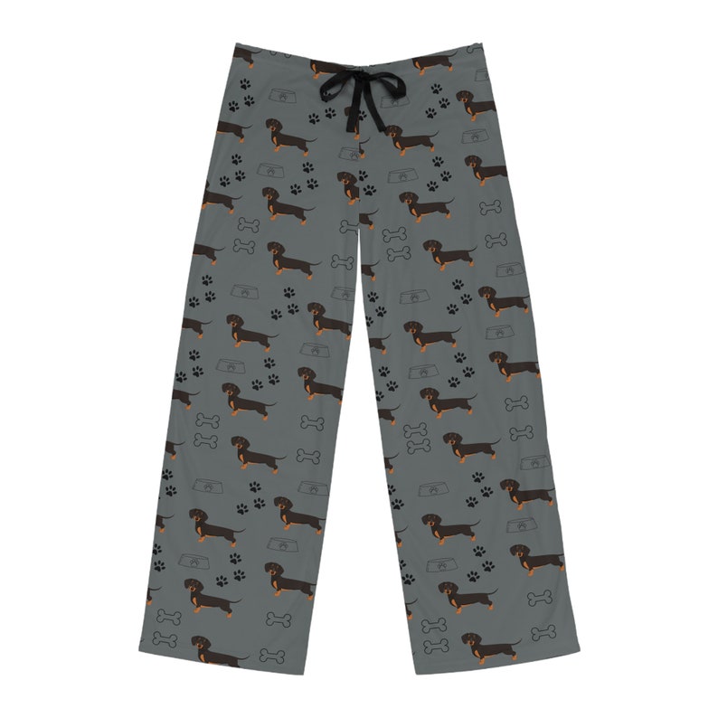 May include: Gray pajama pants featuring an all-over pattern of black and tan dachshund dogs, black paw prints, white dog bones, and gray food bowls. The pants have a black drawstring at the elastic waistband and a straight-leg silhouette.