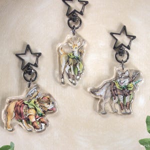 May include: Three clear acrylic keychains with star-shaped clasps. Each keychain features a different animal warrior: a deer, a bulldog, and a wolf, all wearing armor and carrying weapons. The keychains are on a light beige background.