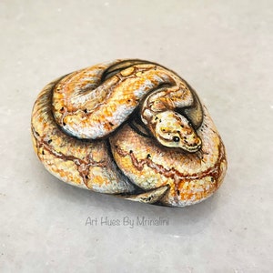 Puede incluir: Una piedra pintada a mano con forma de serpiente enroscada. La serpiente est&aacute; representada en tonos beige, marr&oacute;n y crema, con escamas y marcas detalladas. La obra de arte est&aacute; firmada "Art Hues By Mrinalini".
