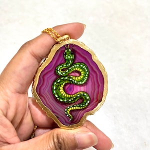 Op de afbeelding: Een groene slang geschilderd op een roze agaat hanger met een gouden rand. De hanger is aan een gouden ketting.