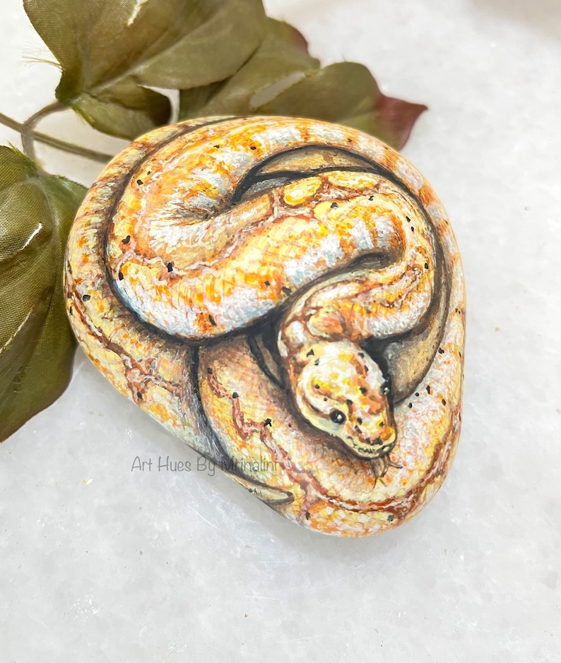Puede incluir: Arte rupestre pintado a mano que representa una serpiente enroscada en tonos amarillos, marrones y blancos. Las escamas de la serpiente est&aacute;n detalladas con motas negras. La obra est&aacute; firmada "Art Hues By Mrinalini".
