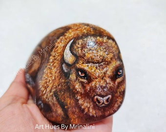 May include: Close-up of a painted rock depicting a brown bison with a white horn. The bison's eyes are painted with a realistic detail. The rock is held in a hand, and the background is a white surface.
