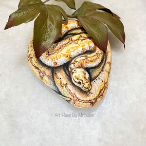 Puede incluir: Una piedra pintada a mano con forma de serpiente enroscada, con escamas detalladas en amarillo, marr&oacute;n y blanco. La obra de arte incluye peque&ntilde;os puntos negros y una cabeza de serpiente con ojos verdes. Se colocan hojas verdes encima. El texto "Art Hues By Mrinalini" es visible.
