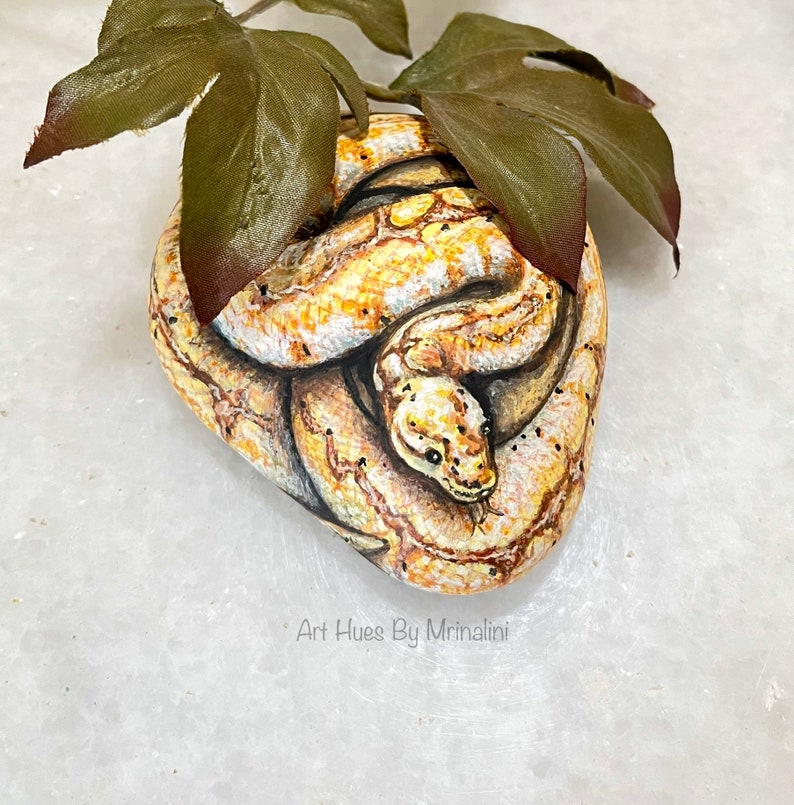 Puede incluir: Una piedra pintada a mano con forma de serpiente enroscada, con escamas detalladas en amarillo, marr&oacute;n y blanco. La obra de arte incluye peque&ntilde;os puntos negros y una cabeza de serpiente con ojos verdes. Se colocan hojas verdes encima. El texto "Art Hues By Mrinalini" es visible.