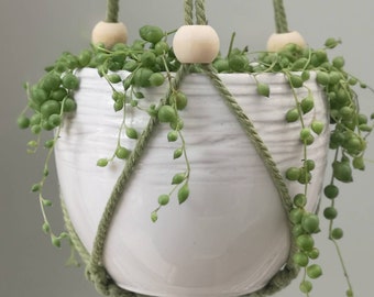 May include: A white ceramic hanging planter with a textured surface, filled with a trailing succulent plant. The planter is suspended by green rope and wooden beads. The plant has small, round, green leaves.