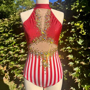 May include: A red and white striped leotard with a halter neckline. The bodice features a sheer panel and is embellished with iridescent rhinestones. The leotard is displayed on a mannequin against a backdrop of greenery.