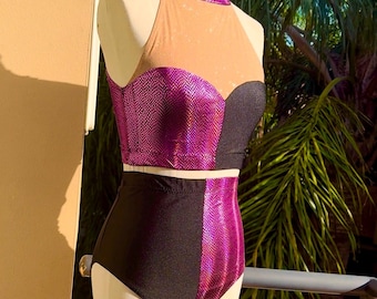 Puede incluir: Un disfraz de baile de dos piezas sobre un maniquí. La parte superior presenta un cuello alto y un panel de malla, con un corpiño morado brillante y negro. Los pantalones de cintura alta son negros con un panel morado. El disfraz se exhibe al aire libre.