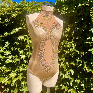 May include: A gold leotard with a high neckline and sheer mesh panel. It is adorned with colourful rhinestones in a decorative pattern. The garment is displayed on a mannequin against a backdrop of green foliage.