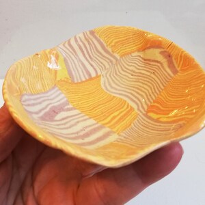 May include: A small, yellow ceramic dish with a wavy rim. The dish is decorated with a patchwork pattern of stripes in shades of yellow, orange, and white.