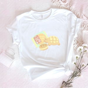 Op de afbeelding: Wit T-shirt met een afbeelding van een schildpad die een gebakje en een pakje "Hokkyokusei" melon pan vasthoudt.