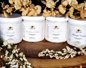 May include: Four white jars of whipped shea butter with different scents: Lavender, Sandalwood Rose, Lemongrass, and Midnight Vanilla. The jars are arranged on a wooden surface with white flowers.
