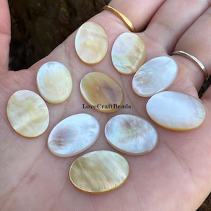 May include: Ten oval-shaped mother of pearl cabochons with iridescent, pearlescent surfaces in shades of white, cream, and pale gold. The smooth, polished stones are arranged in a cluster, and the text LoveCraftBeads is visible in the center.