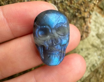 Puede incluir: Una figura de calavera tallada en tonos azules y grises. La calavera está detallada con cuencas de los ojos, una cavidad nasal y dientes. La piedra preciosa tiene una superficie pulida y se sostiene en una mano.