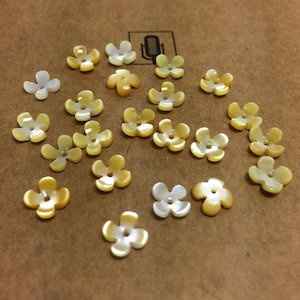 May include: A selection of flower-shaped buttons in yellow and white hues, arranged on a brown surface. Each button features four petals and a central hole for sewing. The buttons display variations in colour and size.