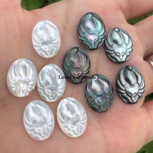 Puede incluir: Una colección de cabujones ovalados, algunos blancos nacarados y otros negros iridiscentes, cada uno tallado con un diseño de cara de zorro. Los cabujones se muestran en una mano, mostrando sus variados colores y texturas. Probablemente son para hacer joyas.