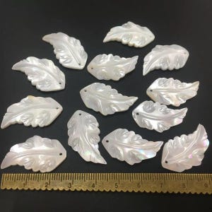 Può includere: Una collezione di ciondoli bianchi iridescenti a forma di foglia. Ogni ciondolo presenta venature dettagliate e un piccolo foro per l'infilatura. Le foglie sono disposte su una superficie scura, con un righello in basso.