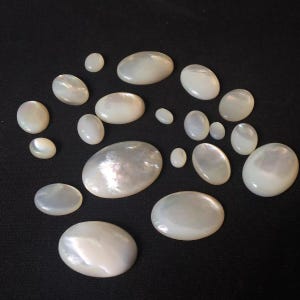 May include: A collection of various oval-shaped mother-of-pearl cabochons in different sizes. Each piece features a smooth, polished surface with a natural iridescent white sheen, displaying subtle variations in lustre and light reflection.
