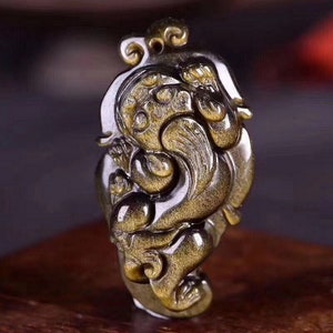 May include: A gold and black carved pendant featuring a mythical creature with intricate details. The pendant is made of tiger's eye stone and has a shiny, polished finish.