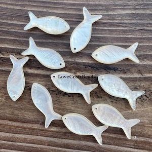 Może przedstawiać: Kolekcja iryzujących koralików w kształcie rybki. Koraliki są perłowo-białe z subtelnymi odcieniami i fakturą. Każda rybka ma mały otwór do nawlekania. Koraliki ułożone są na drewnianej powierzchni.