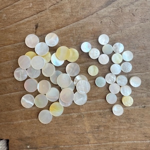 Può includere: Una collezione di dischi rotondi in madreperla bianca e iridescente. I dischi sono di varie dimensioni e sono sparsi su una superficie di legno.