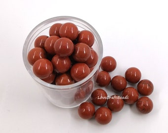 May include: A clear plastic container filled with red jasper beads. The beads are round and smooth, and they are spilling out of the container onto a white surface.  "LoveCraftBeads" is written on the white surface.