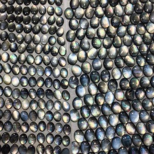 Peut inclure: Vue aérienne d'une collection de pierres précieuses ovales polies. Les pierres présentent une gamme de couleurs, notamment le gris, le noir et des teintes irisées. Les pierres sont disposées de manière rapprochée, créant un motif dense.