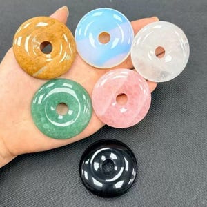 Puede incluir: Colección de siete colgantes de piedras preciosas en forma de donut de varios colores, incluyendo marrón, azul, blanco, verde, rosa y negro. Cada colgante tiene forma circular con un agujero en el centro. Las piedras son lisas y pulidas.