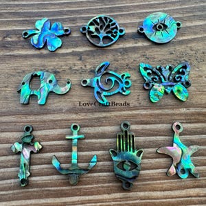 Puede incluir: Una colección de dijes iridiscentes de concha de abulón en varias formas, incluyendo una flor, árbol, ojo, elefante, tortuga, mariposa, cruz, ancla, mano de Hamsa y estrella de mar. Los dijes tienen una paleta de colores naturales y brillantes de azules, verdes y morados.